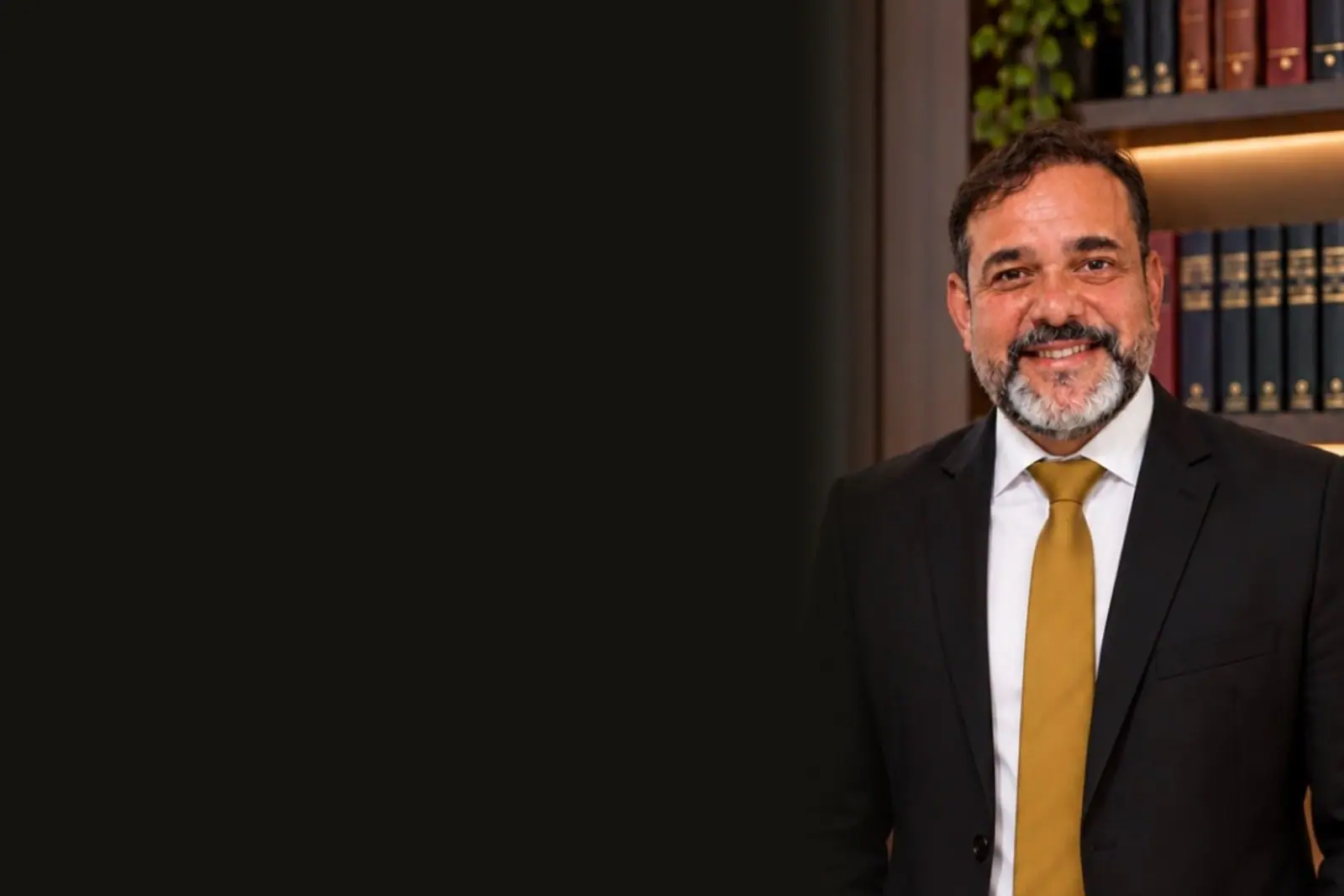 Everson Motta de terno escuro e gravata dourada, sorrindo, diante da estante de livros do escritório